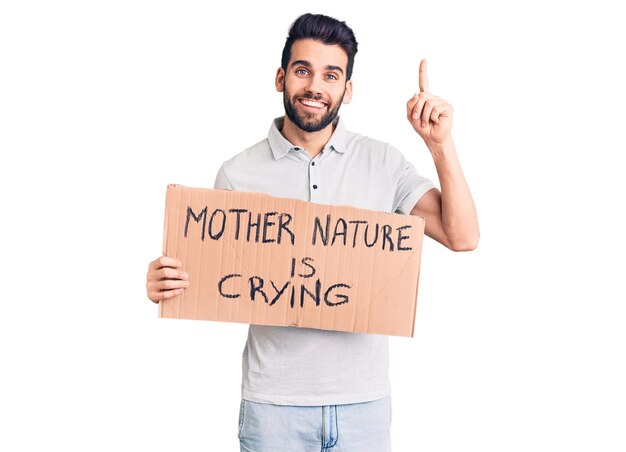 Jeune bel homme avec barbe tenant mère nature pleure bannière en carton surpris par une idée ou une question pointant du doigt avec un visage heureux, numéro un