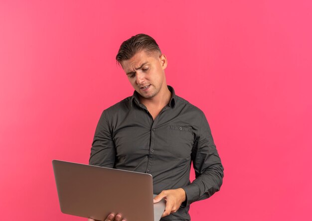 Jeune bel homme blond agacé tient et regarde un ordinateur portable isolé sur fond rose avec espace de copie