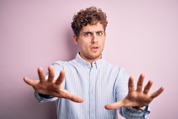 Photo gratuite jeune bel homme blond aux cheveux bouclés portant une chemise rayée sur fond blanc faisant un geste d'arrêt avec les mains paumes expression de colère et de frustration