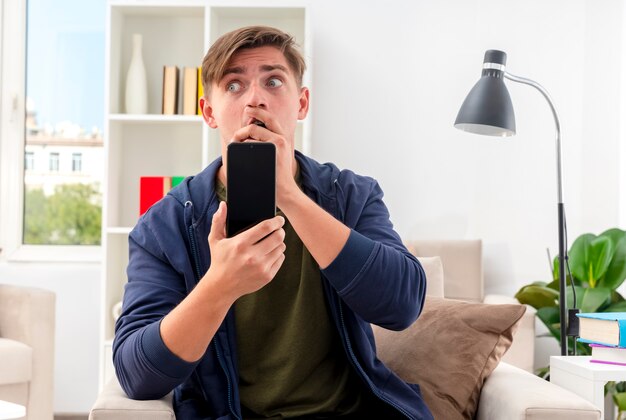 Jeune bel homme blond choqué est assis sur un fauteuil, mettant la main sur la bouche