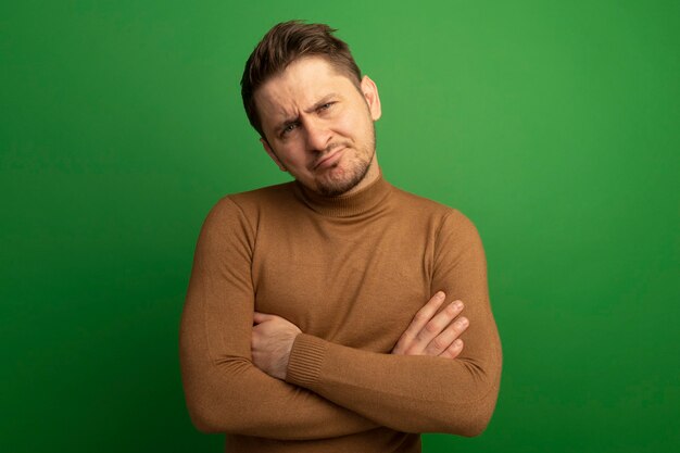 Jeune bel homme blond confus debout avec une posture fermée à la recherche d'isolement sur un mur vert avec espace de copie