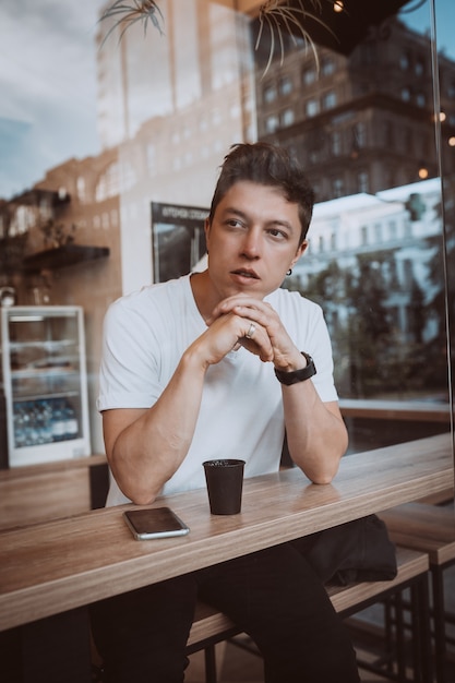 Un jeune et bel homme boit son café du matin dans un café. Photo derrière une vitre