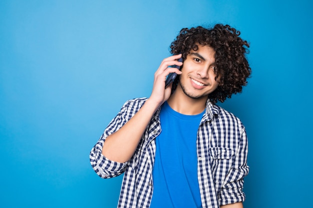 Jeune bel homme bouclé parler au téléphone isolé sur mur bleu