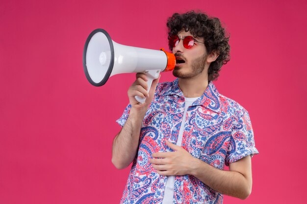 Jeune bel homme bouclé portant des lunettes de soleil parlant par le haut-parleur avec la main sur son ventre sur un mur rose isolé avec copie espace