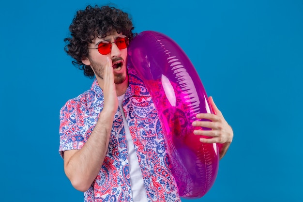 Jeune Bel Homme Bouclé Portant Des Lunettes De Soleil Tenant Un Anneau De Bain Criant à Quelqu'un Avec La Main Près De La Bouche Sur Un Mur Bleu Isolé Avec Copie