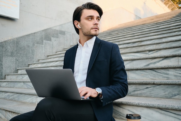 Jeune bel homme brune barbu en chemise blanche et costume classique assis dans les escaliers en plein air travaillant sur un ordinateur portable tout en regardant rêveusement de côté