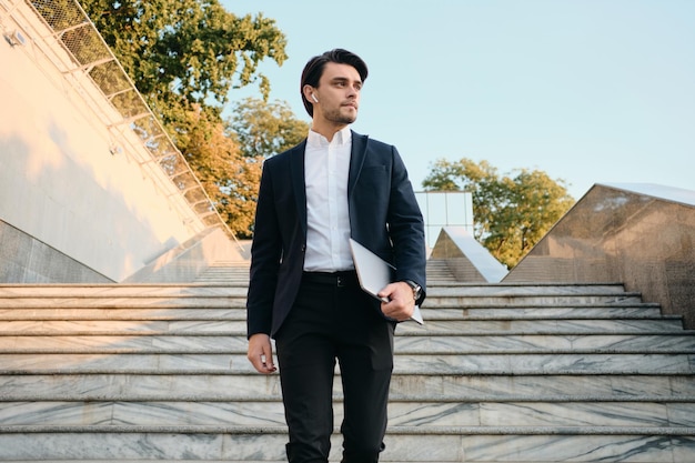 Jeune bel homme brune barbu en chemise blanche et costume classique avec des écouteurs sans fil tenant un ordinateur portable à la main regardant rêveusement de côté en plein air