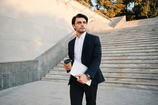 Jeune bel homme brune barbu en chemise blanche et costume classique avec des écouteurs sans fil tenant une tasse de café à emporter et un ordinateur portable dans les mains regardant pensivement de côté dans le parc de la ville