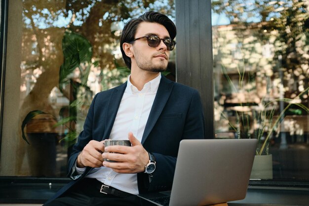 Jeune bel homme brune barbu en chemise blanche, veste classique et lunettes de soleil regardant pensivement de côté tenant une tasse de café dans les mains assis avec un ordinateur portable seul dans la rue