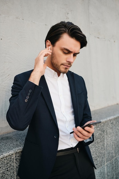 Jeune bel homme brune barbu sérieux en chemise blanche et costume classique avec des écouteurs sans fil utilisant judicieusement le téléphone portable tout en se tenant près du mur gris dans la rue
