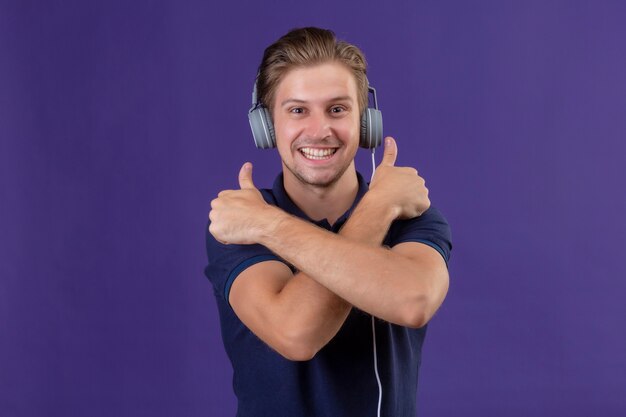 Jeune bel homme avec un casque debout avec les bras croisés montrant les pouces vers le haut souriant joyeusement sur fond violet