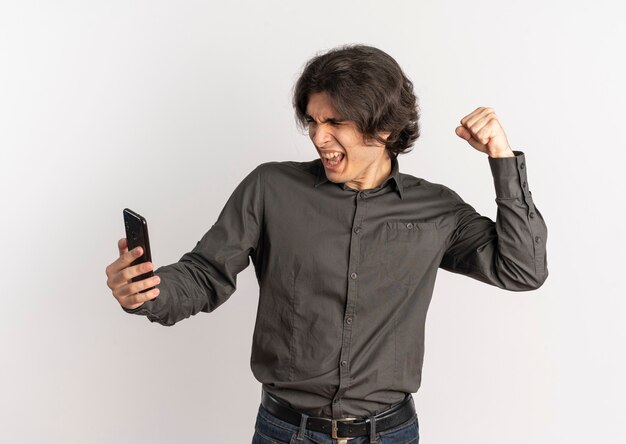 Jeune bel homme caucasien agacé lève le poing et regarde le téléphone isolé sur fond blanc avec espace de copie