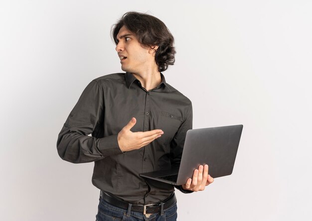 Jeune bel homme caucasien agacé tient et pointe à l'ordinateur portable à côté isolé sur fond blanc avec copie espace