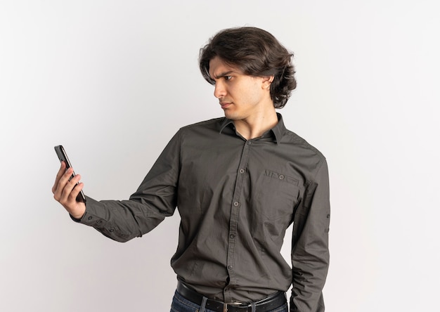 Jeune bel homme caucasien agacé tient et regarde le téléphone isolé sur fond blanc avec copie espace