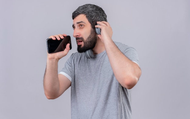 Jeune bel homme caucasien portant des écouteurs faire semblant de chanter et d'utiliser son téléphone portable comme microphone et regardant de côté avec la main sur le casque isolé sur fond blanc avec espace de copie