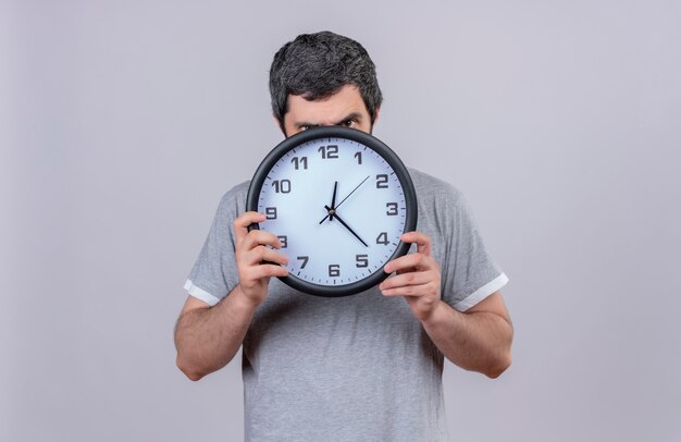 Jeune bel homme caucasien tenant et regardant la caméra par derrière l'horloge isolé sur fond blanc avec copie espace