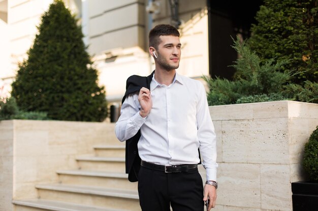 Jeune bel homme en chemise blanche avec des écouteurs sans fil tenant une veste noire sur l'épaule tout en regardant rêveusement de côté en plein air