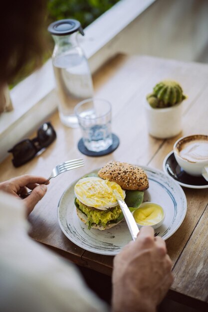 Jeune bel homme en chemise blanche ouverte, prenant son petit déjeuner dans un café avec un hamburger végétarien, buvant du café, mode de vie dans une île tropicale, vie à Bali.