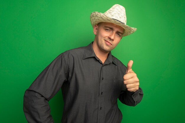 Jeune bel homme en chemise grise et chapeau d'été à l'avant, souriant avec un visage heureux et montrant les pouces vers le haut debout sur le mur vert