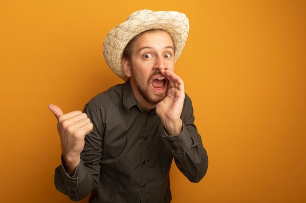 Jeune bel homme en chemise grise et chapeau d'été criant ou appelant quelqu'un avec la main près de la bouche et pointant avec l'index sur quelque chose