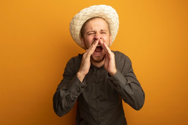 Jeune bel homme en chemise grise et chapeau d'été criant ou appelant quelqu'un avec les mains près de la bouche excité et émotionnel