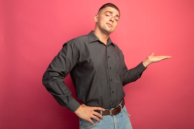 Jeune bel homme en chemise grise présentant quelque chose avec le bras de sa main à la confiance debout sur le mur rose
