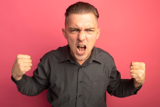 Jeune bel homme en chemise grise serrant les poings en criant fou fou et en colère debout sur le mur rose