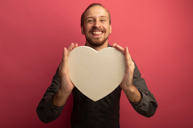 Jeune bel homme en chemise grise tenant coeur en carton avec grand sourire sur le visage