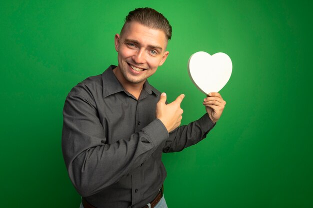 Jeune bel homme en chemise grise tenant coeur en carton pointant avec l'index sur elle souriant avec visage heureux debout sur mur vert
