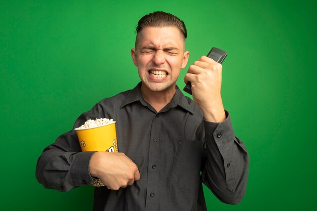 Jeune bel homme en chemise grise tenant un seau avec du pop-corn à l'aide de la télécommande tv hurlant fou fou et frustré debout sur le mur vert