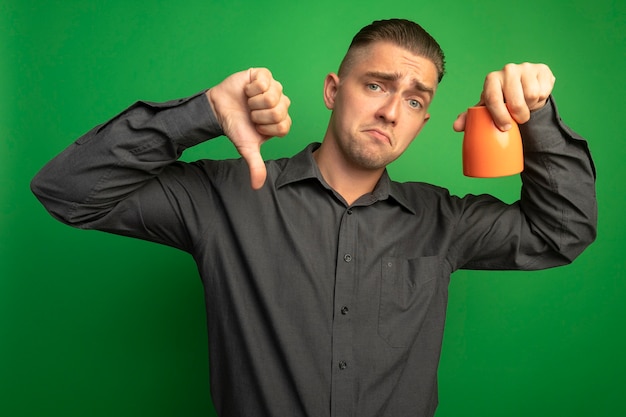 Jeune bel homme en chemise grise tenant une tasse orange à l'avant mécontent montrant les pouces vers le bas debout sur un mur vert