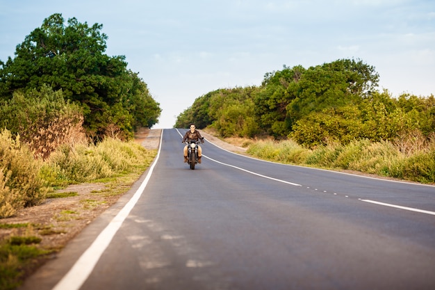 Jeune bel homme à cheval sur la moto à la route de campagne.