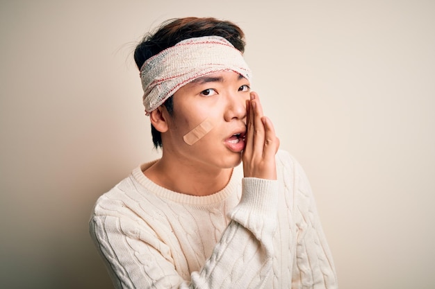 Photo gratuite jeune bel homme chinois blessé dans un accident portant un bandage et des bandes sur la tête main sur la bouche racontant une rumeur secrète chuchotant une conversation malveillante