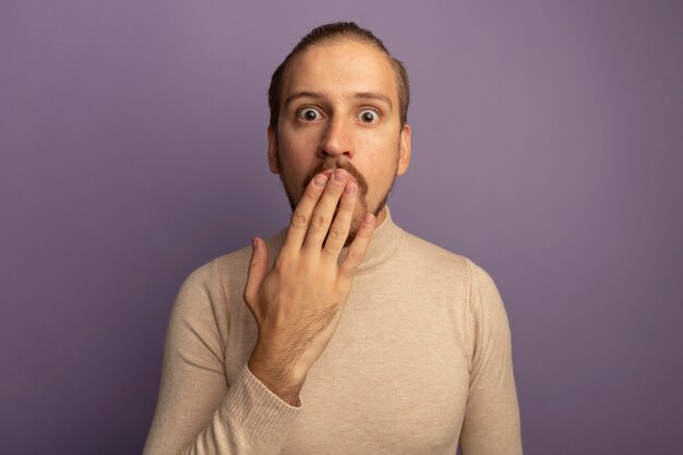 Jeune bel homme en col roulé beige à l'avant d'être choqué couvrant la bouche avec la main debout sur le mur lilas