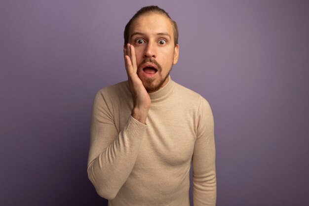 Jeune bel homme en col roulé beige à l'avant avec la main ner bouche étant surpris debout sur mur lilas