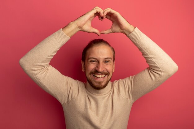 Jeune bel homme en col roulé beige faisant le geste du cœur avec les doigts sur sa tête souriant joyeusement