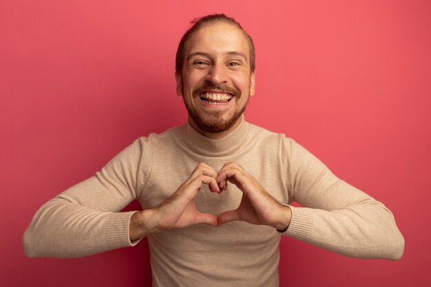 Jeune bel homme en col roulé beige faisant le geste du coeur sur la poitrine heureux et joyeux souriant debout sur le mur rose