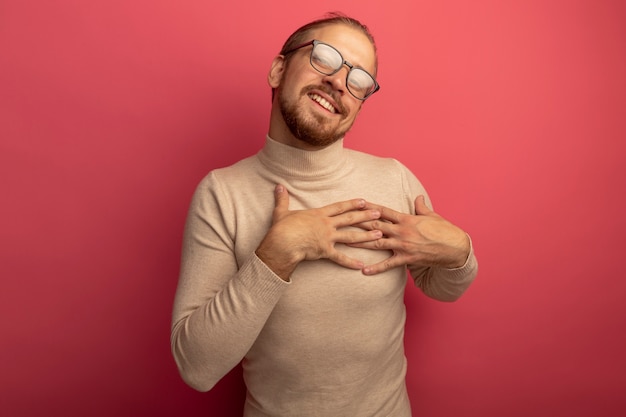 Jeune bel homme en col roulé beige et lunettes souriant joyeusement tenant les mains sur sa poitrine se sentant reconnaissant debout sur le mur rose