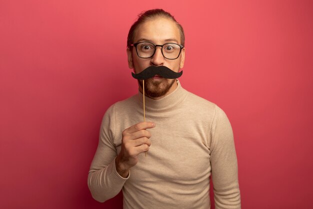 Jeune bel homme en col roulé beige et lunettes tenant une drôle de moustache sur bâton debout sur un mur rose