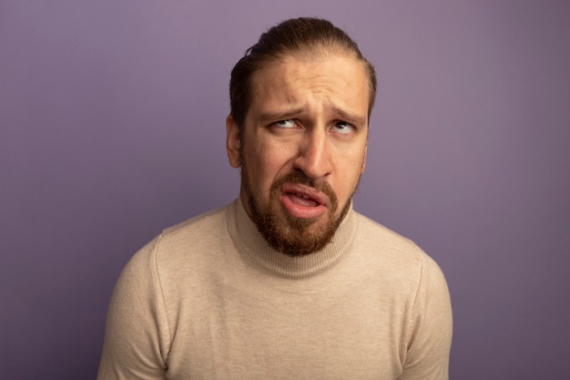 Jeune bel homme en col roulé beige à la recherche avec une expression triste debout sur un mur lilas