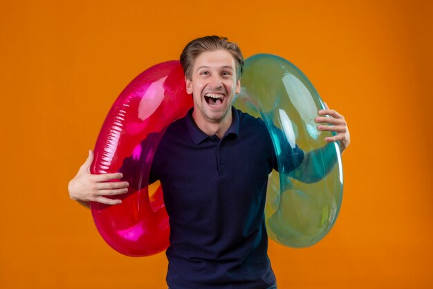 Jeune bel homme debout avec des anneaux gonflables surpris et heureux avec la bouche grande ouverte sur fond orange