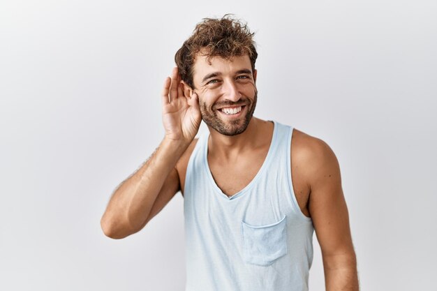 Jeune bel homme debout sur fond isolé souriant avec la main sur l'oreille en écoutant une rumeur ou des commérages. notion de surdité.