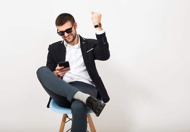 Jeune bel homme élégant hipster en veste noire, style affaires, chemise blanche, isolé, assis détendu sur une chaise de bureau, à l'aide de smartphone, souriant, heureux, positif, lunettes de soleil, geste, émotionnel