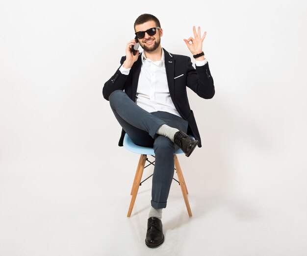 Jeune bel homme élégant hipster en veste noire, style affaires, chemise blanche, isolé, assis détendu sur une chaise de bureau, parler sur smartphone, souriant, heureux, positif, lunettes de soleil