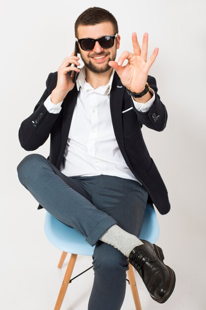 Jeune bel homme élégant hipster en veste noire, style affaires, chemise blanche, isolé, assis détendu sur une chaise de bureau, parler sur smartphone, souriant, heureux, positif, lunettes de soleil