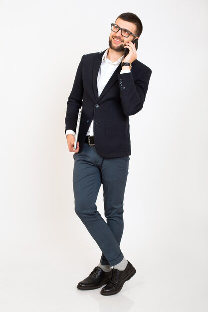 Jeune bel homme élégant hipster en veste noire, style affaires, chemise blanche, isolé, fond blanc, souriant, debout pleine hauteur, à la recherche de confiance, tenant un ordinateur portable, parler sur smartphone