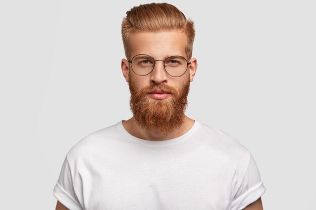 Jeune bel homme hipster a une barbe et une moustache épaisses au gingembre, une coupe de cheveux à la mode, vous regarde sérieusement
