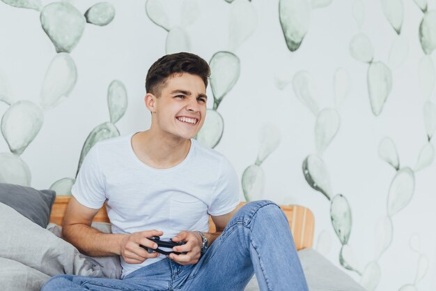 Le jeune bel homme sur le lit dans sa chambre joue à un jeu avec un joystick