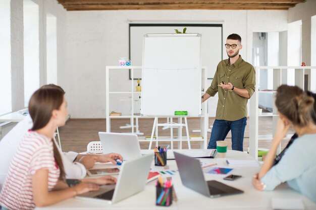 Jeune bel homme en lunettes et chemise debout près du tableau tout en discutant d'un nouveau projet avec des collègues Groupe de personnes travaillant ensemble de manière réfléchie dans un bureau moderne