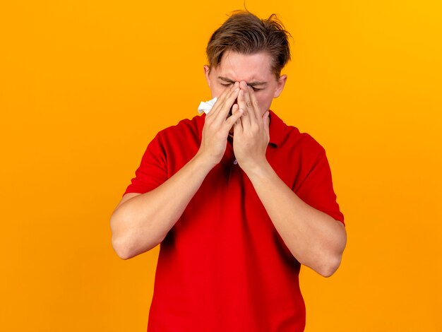 Jeune bel homme malade blonde tenant la serviette en gardant les mains sur le visage avec les yeux fermés isolé sur un mur orange avec espace de copie